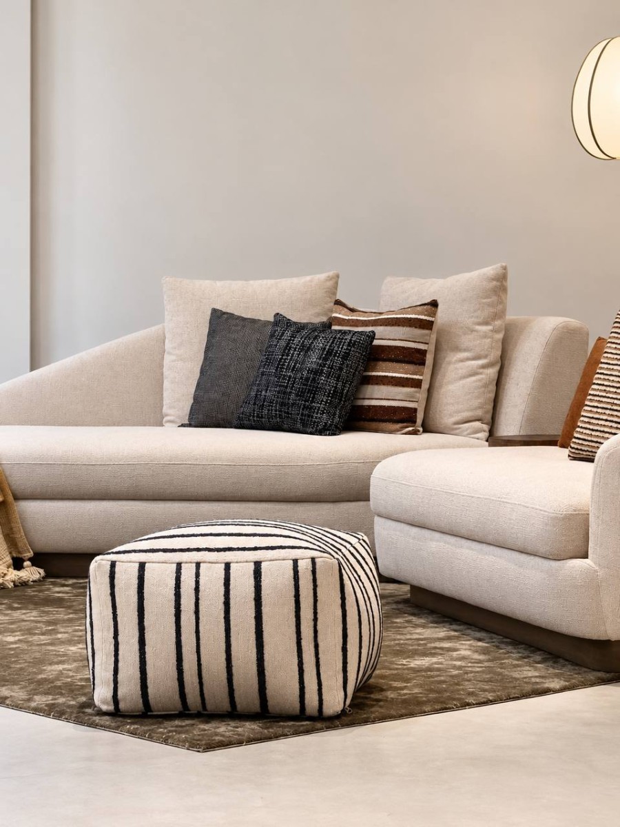 Black and cream striped pouf by NicChris styled in a modern living room with a curved cream sofa, wooden lounge chair, and olive green rug